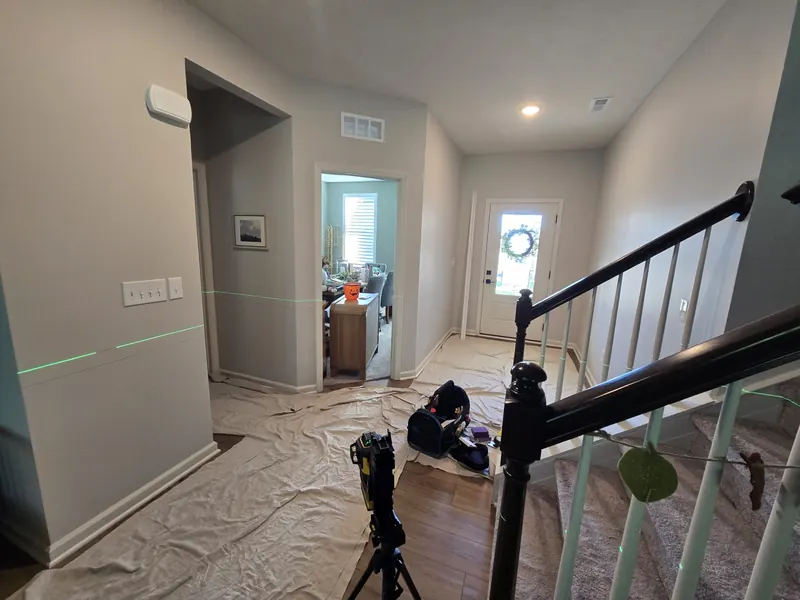 Foyer and staircase during box moulding installation, a green laser level line across the bare gray wall with a tripod and drop cloths on the floor