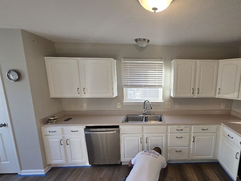 Kitchen before cabinet painting, white raised-panel cabinets with silver pulls around a double sink and stainless dishwasher, a painter kneeling at the base cabinets