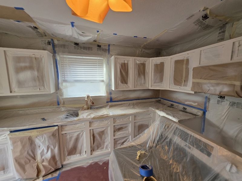 Same kitchen masked off in plastic sheeting and paper, cabinet boxes sanded and primed white with door openings covered, ready for paint