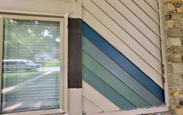 Close-up of a residential window with blue and green paint swatches applied to diagonal wood siding during color sampling.