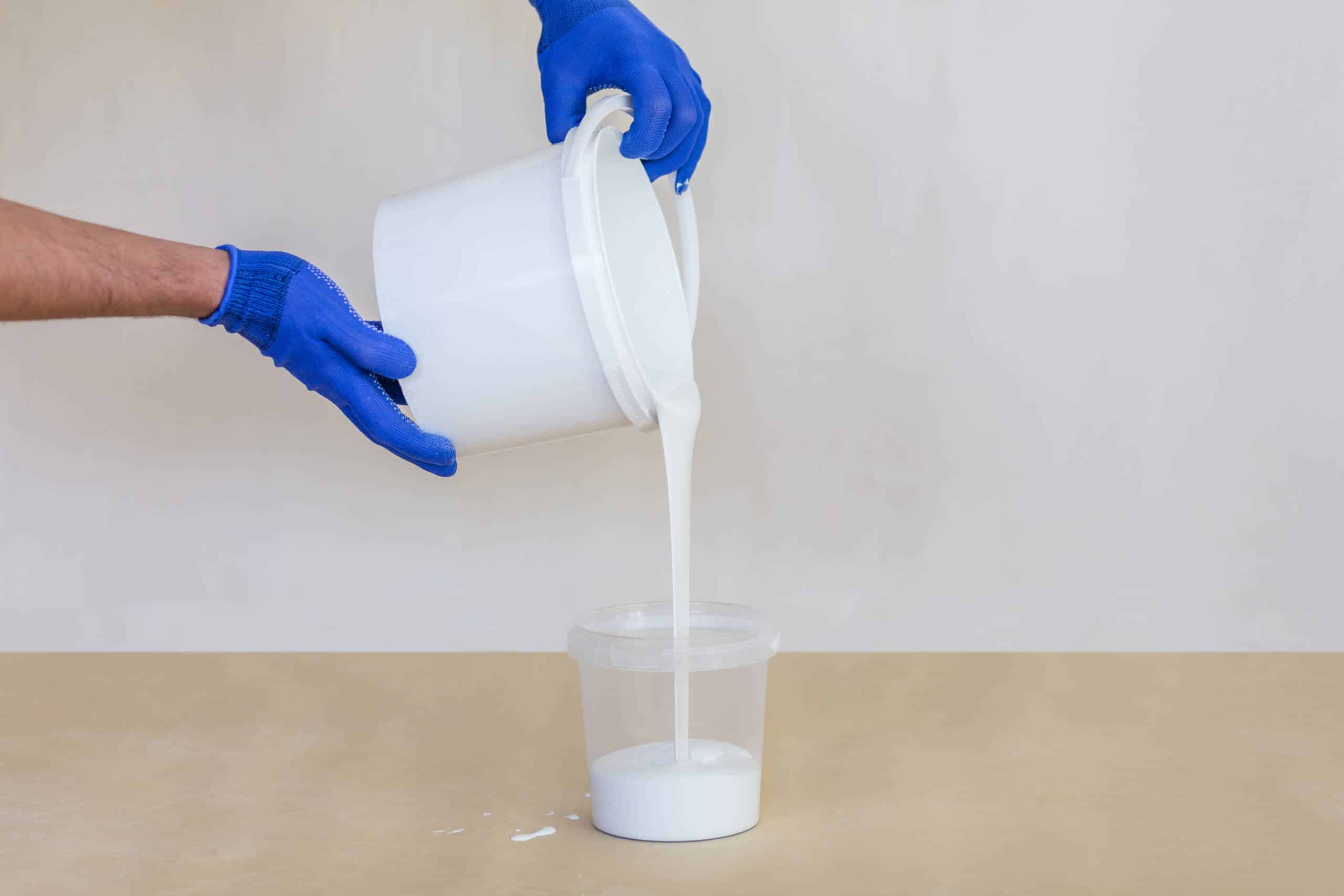 A pair of hands wearing blue work gloves carefully pouring thick white paint from a large bucket into a smaller clear container. This demonstration of professional preparation highlights the precision and care involved in painting services indianapolis in geist. Guy painting utilizes high-quality materials and expert techniques to deliver a clean, professional finish for every residential painting project.